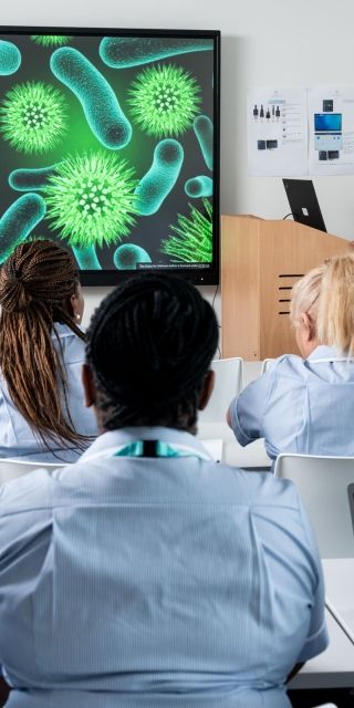The back of the heads of Nursing students in uniform sat in a classroom facing a teacher at the front of the class 