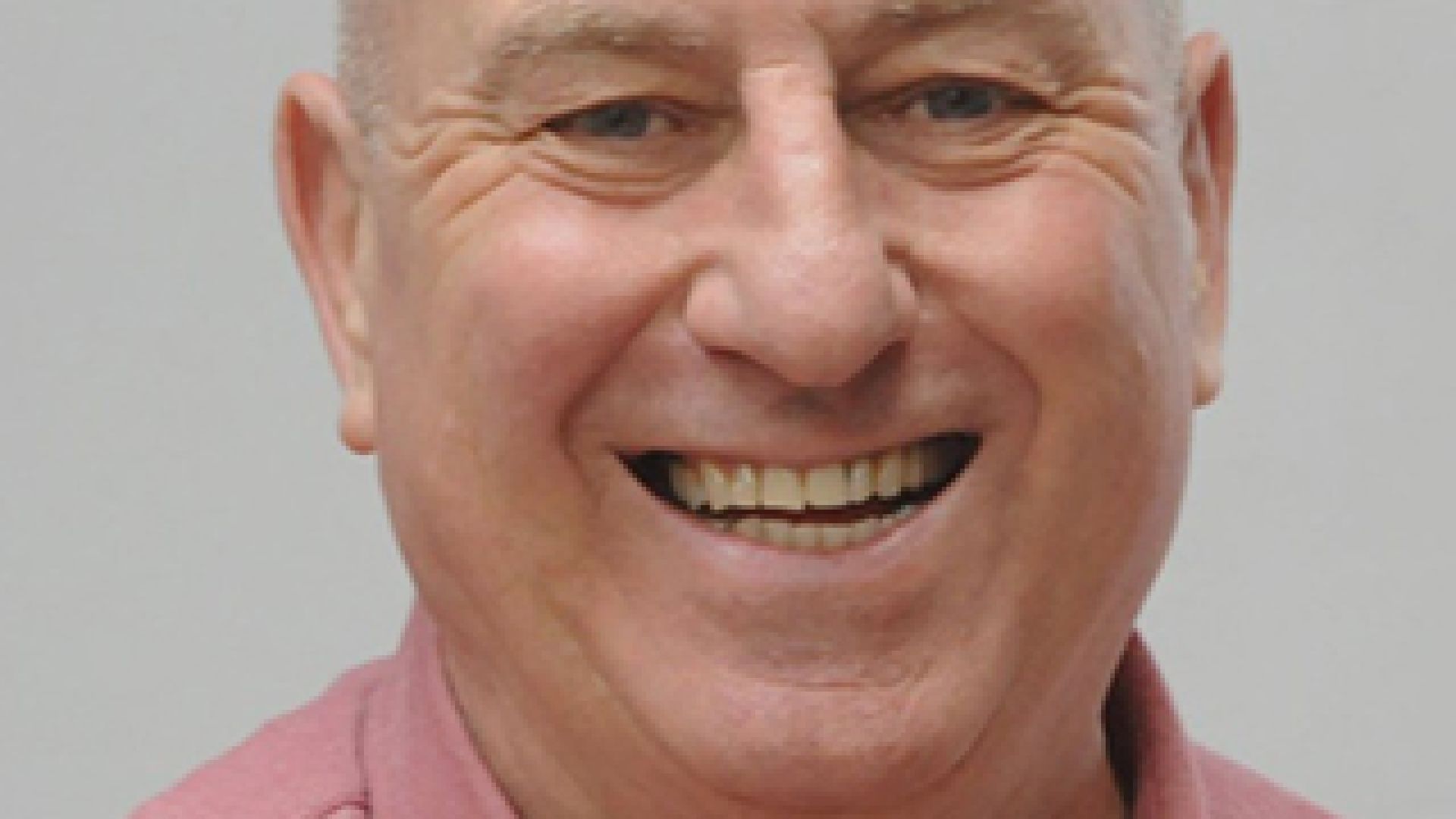 Head and shoulders shot of a smiling Richard Jones wearing a pink shirt stood in front of a grey wall looking into the camera