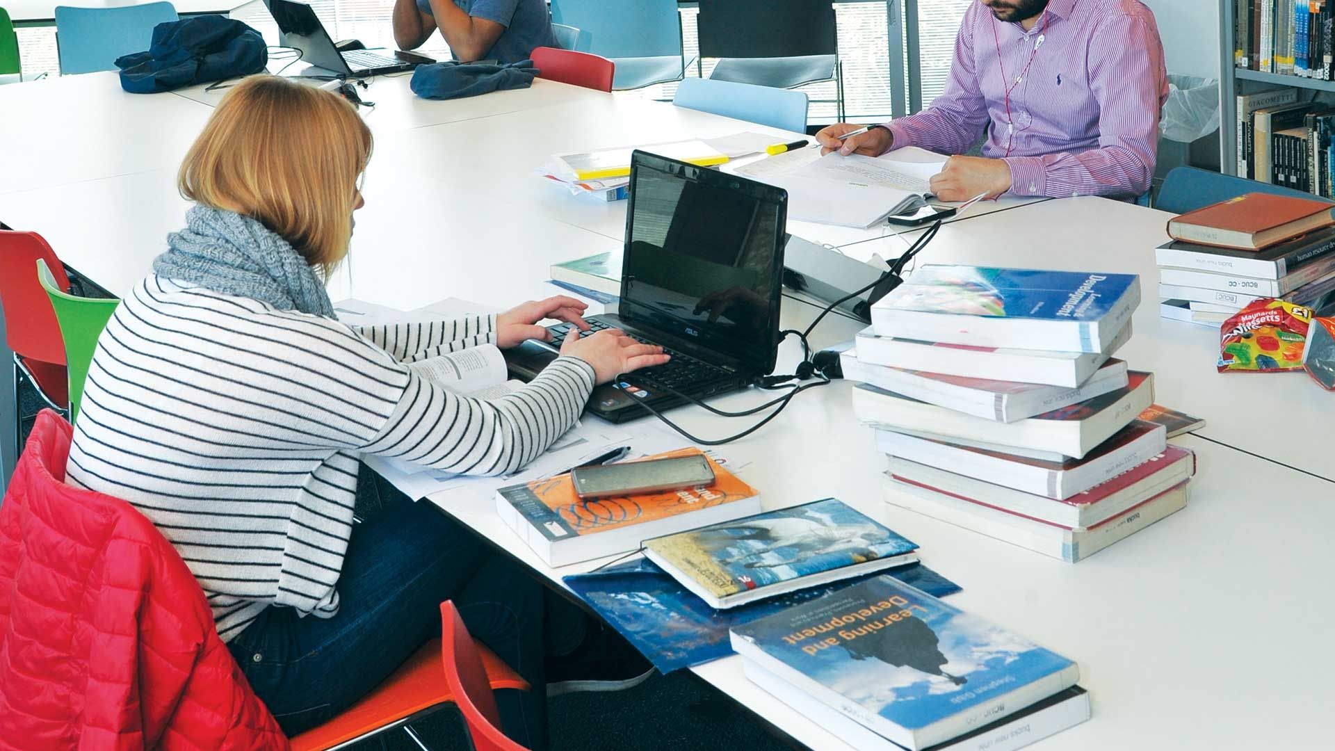 BNU female student in library with lots pf books round her and laptop