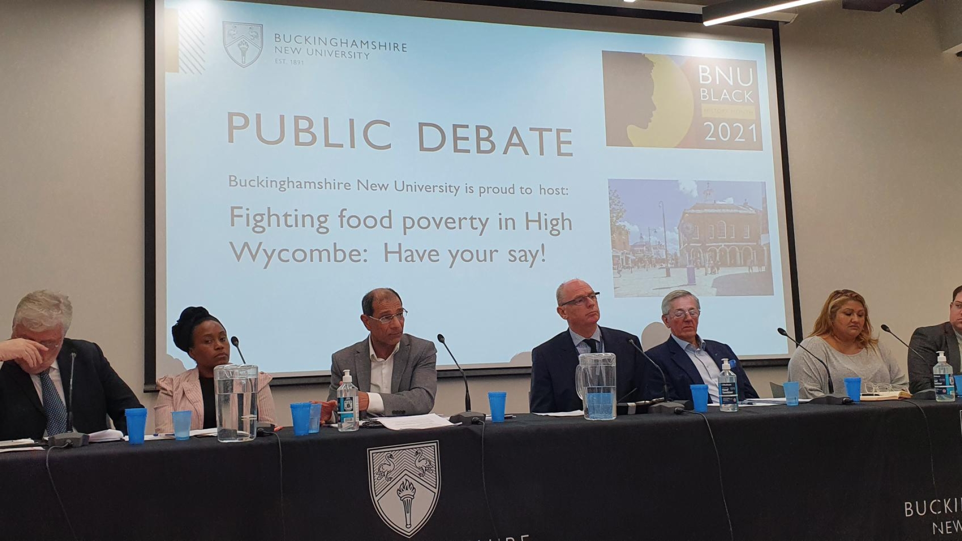 7 panellists sit at table at public debate