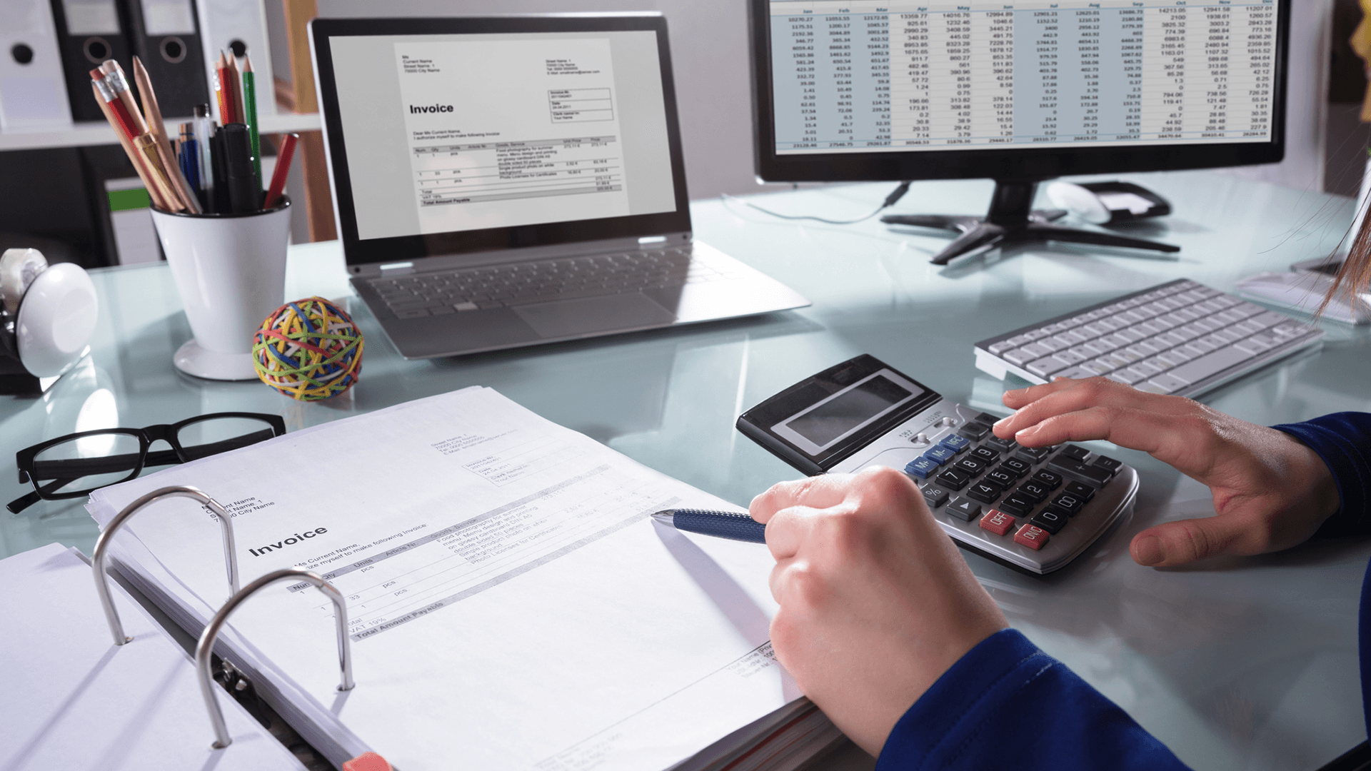 An accounting and finance student sat at a desk looking at invoice information on two computer screens with a calculator