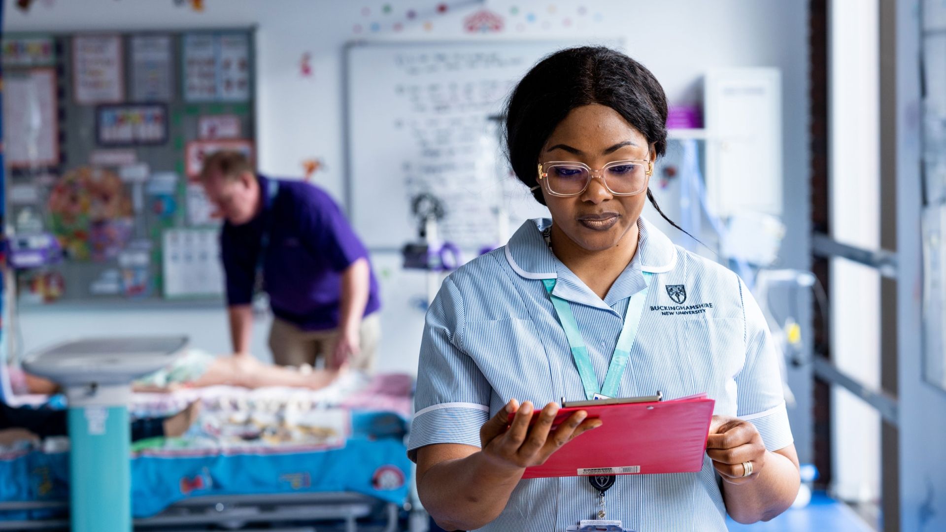 A student nurse in uniform stood in a simulation ward looking down at a tablet screen