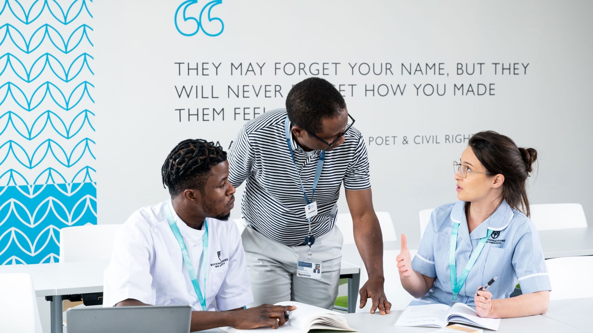 Two nursing students sat at a desk in a class with the teacher stood in between them in discussion