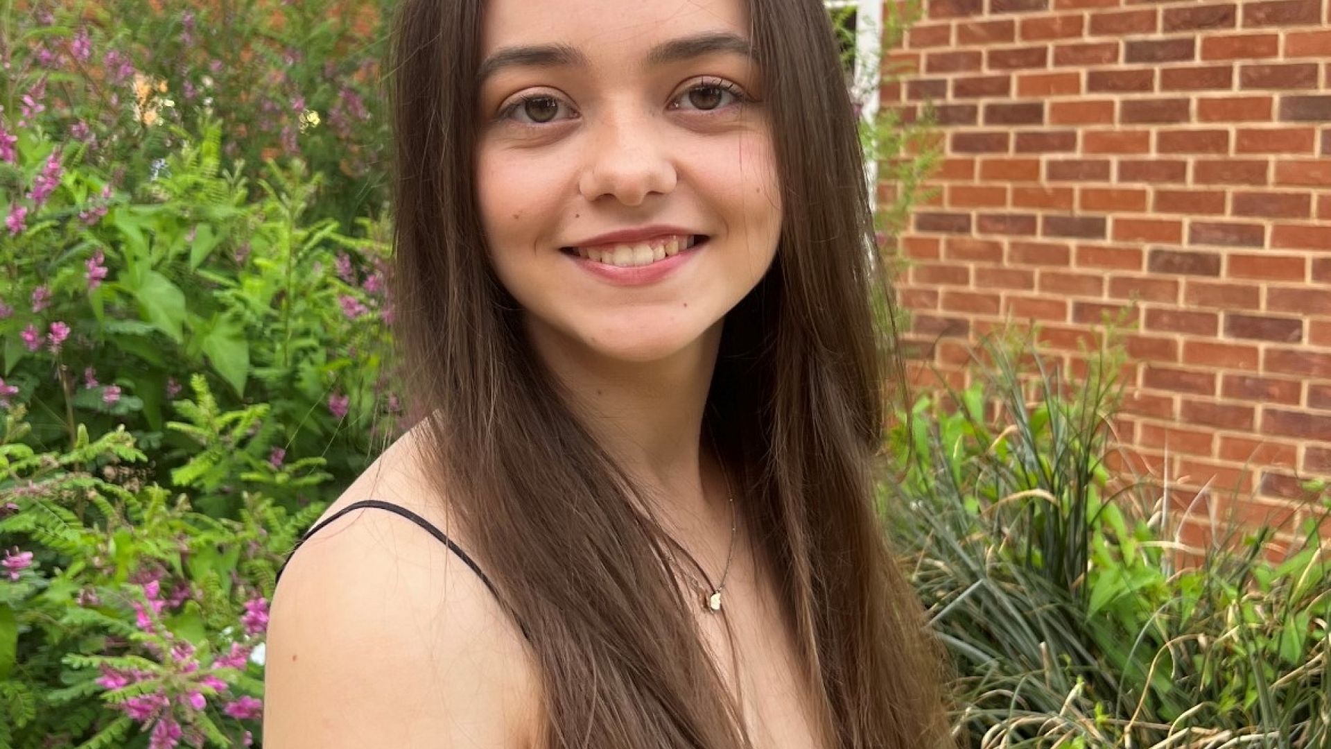 Student Sophie, who has pale skin and waist-length brown hair, stands in front of a brick building.
