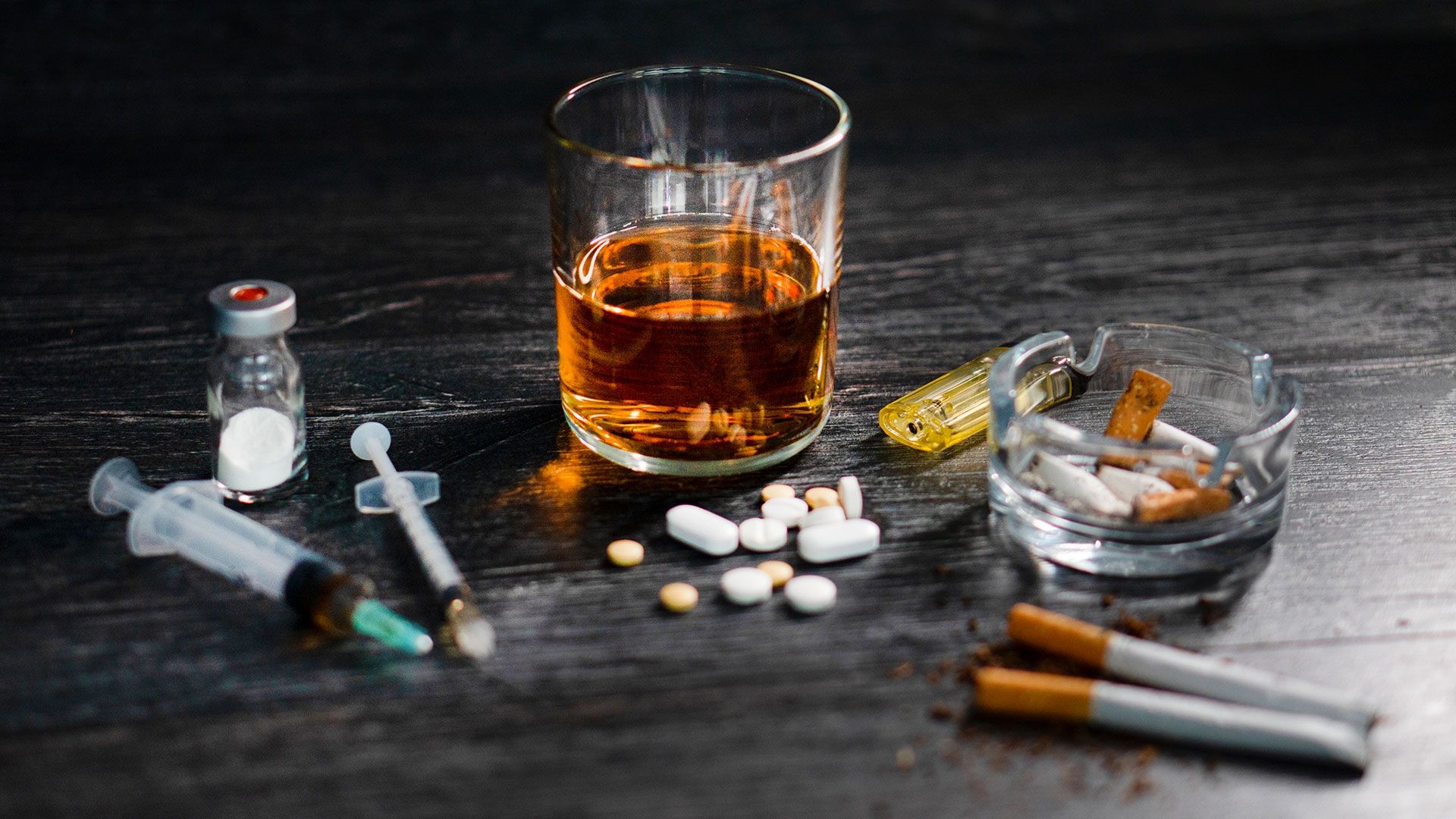glass of whiskey, ashtray, cigarettes, pills and drug paraphernalia on a table