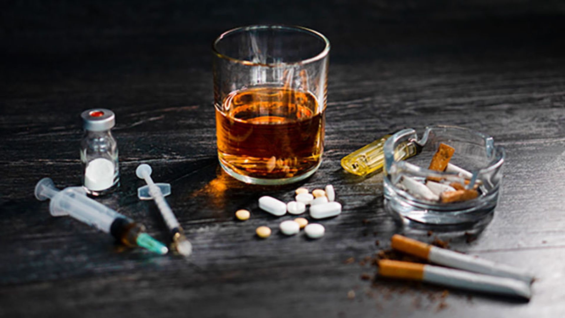 glass of whiskey, ashtray, cigarettes, pills and drug paraphernalia on a table