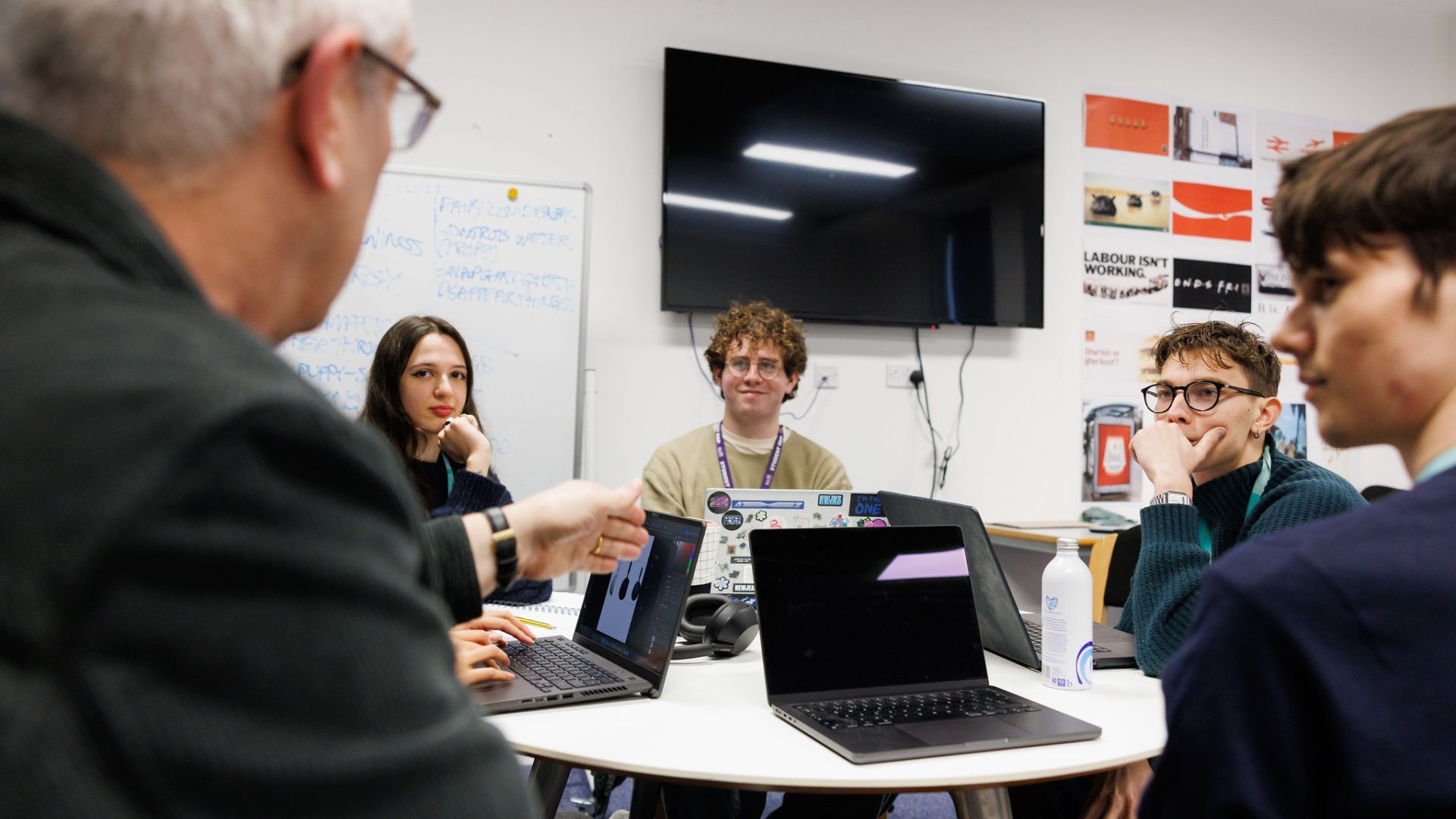 Students sitting around a table with their laptops, talking to a lecturer