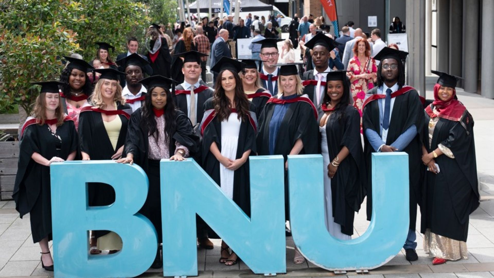 students in graduation gowns and caps standing in front of a BNU sign smiling and waving