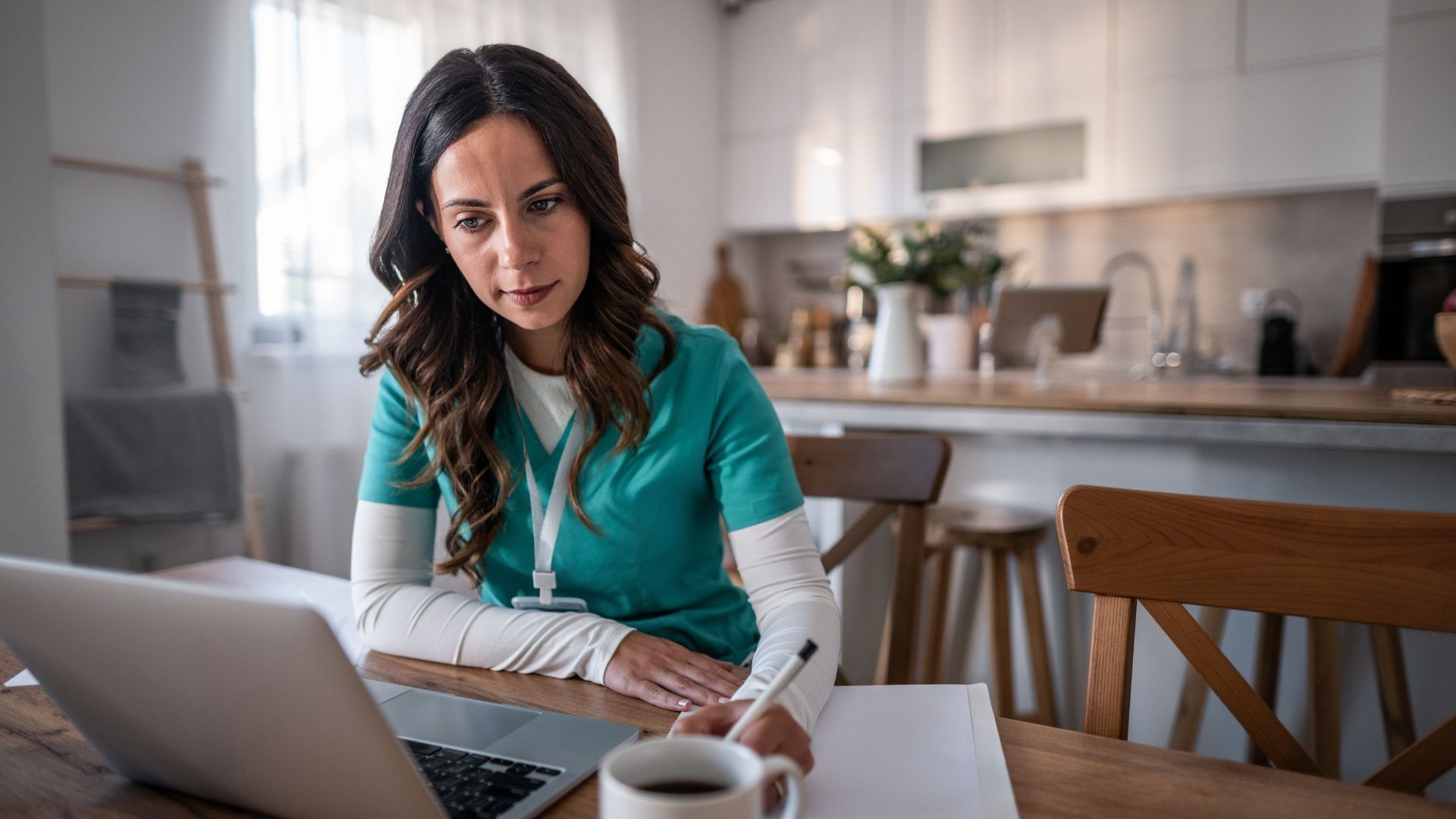 A student nurse sat in a kitchen at a table making notes whilst looking at a laptop screen.