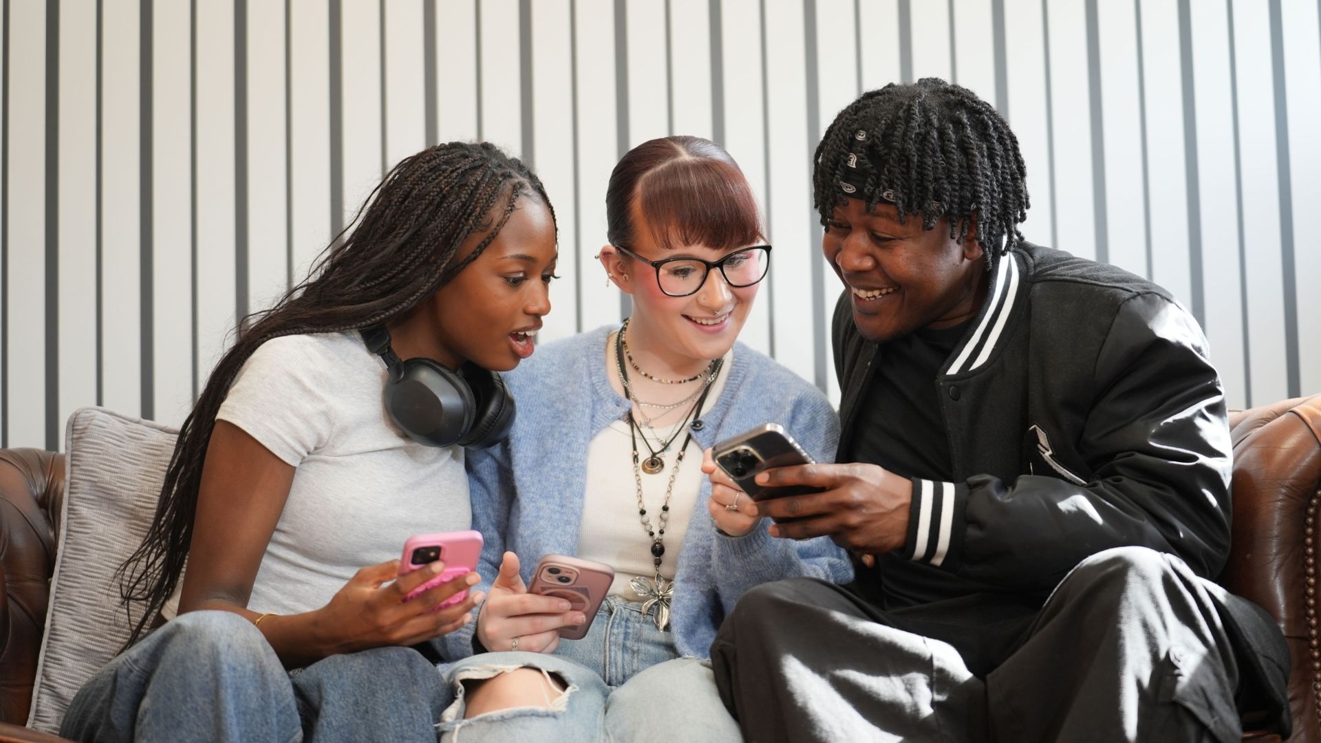 Three young people sit together on a leather sofa, smiling and reacting with surprise as they look at a smartphone held by one of them against a backdrop of vertical black and white stripes.
