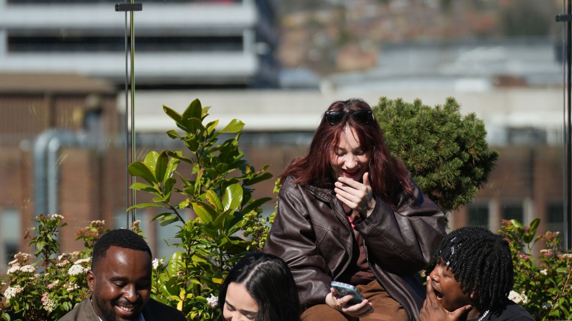  Four friends sit on an outdoor wooden bench and laugh together while looking at their smartphones on a sunny day with a cityscape in the background.