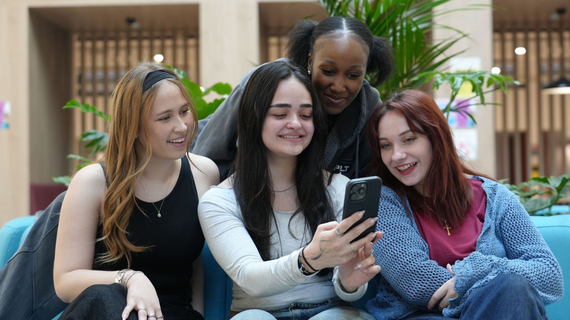 Four young women are gathered together on a blue sofa in a bright indoor space, smiling as they look at a smartphone being held by the person in the center.