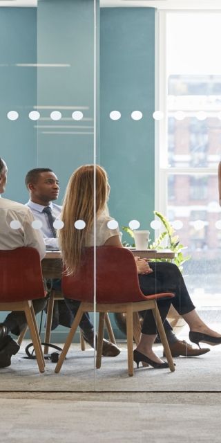 A lady stood up showing a presentation with 6 others sat at a desk watching
