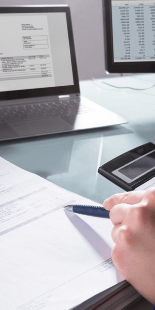 An accounting and finance student sat at a desk looking at invoice information on two computer screens with a calculator