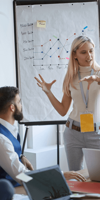 A woman stood in front of a white board talking to 5 adults all sat at a table looking in her direction