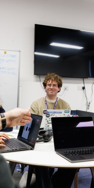 Students sitting around a table with their laptops, talking to a lecturer
