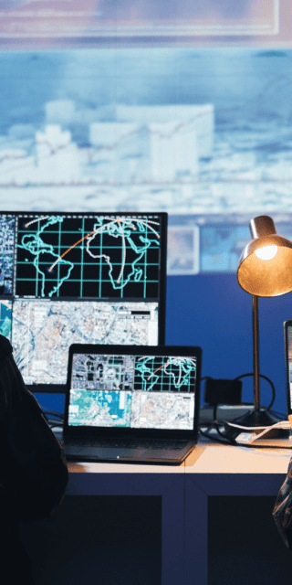 Two people sat at a desk wearing headphones looking at numerous computer screens with lots of data on.