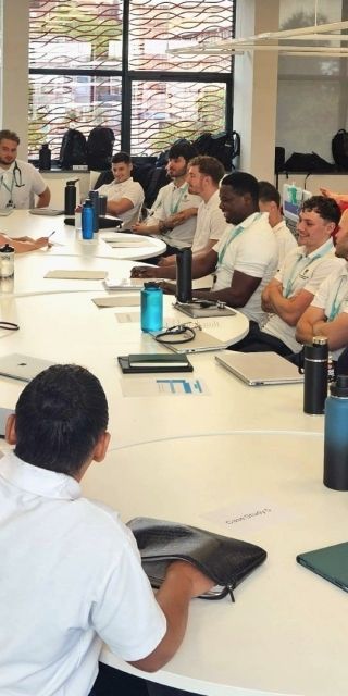 A class of Physiotherapy students sat round desks as a lecturer is stood on the left talking to students.