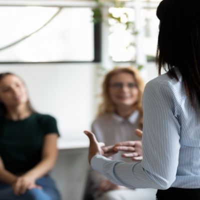A woman with her back to the camera delivers a speech to three other females