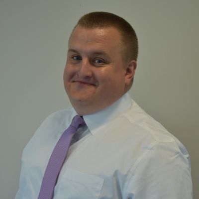 A head and shoulders shot of a smiling Danny Clarke standing side on looking into the camera wearing a shirt and tie