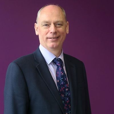 A top half shot of Professor Nick Braisby wearing a black suit stood against a purple background 