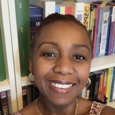A head shot of a smiling Sena Quaye looking directly into the camera stood against a book shelf