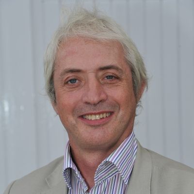 A head and shoulders shot of Ian Chisolm Bunting wearing a beige suit jacket with a grey, red, and white striped shirt stood face on looking directly into the camera