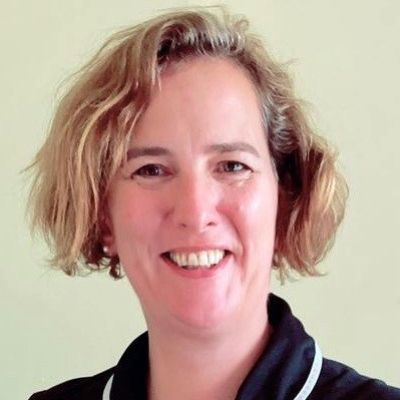 A head and shoulders shot of nurse Natasha Shand wearing a navy blue nursing uniform stood in front of a grey wall looking into the camera
