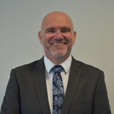 A top half shot of a smiling Shane Roadnight wearing a grey suit jacket, white shirt and a blue floral tie looking directly into the camera
