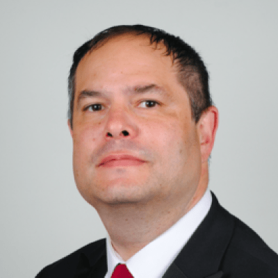 Head shot of a smiling Ze'ev Portner wearing a black suit jacket, white shirt and red tie looking directly into the camera and stood face on