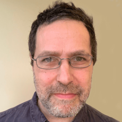 Head shot of Alan Green wearing glasses stood against a light brown wall looking directly into the camera and stood face on