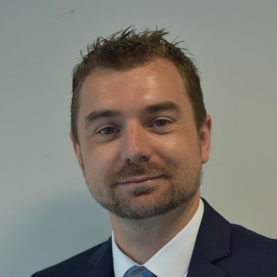 Head and shoulders shot of a smiling James Ellis wearing a navy blue suit jacket with a white shirt and blue tie stood in front of a grey wall