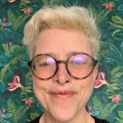 Head shot of a smiling Keelin Howard wearing glasses stood facing straight on into the camera in front of a green floral wall