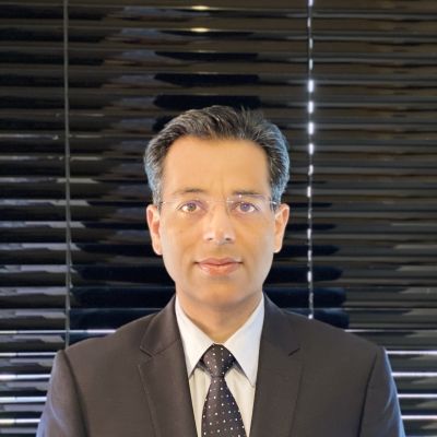 Head and shoulders photo of a smiling Naveed Kapadia wearing a black suit jacket, white shirt and black tie stood facing directly into the camera