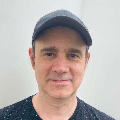 Head and shoulders shot of a smiling Paul Martin wearing a cap with a blue t shirt on stood in front of a white wall