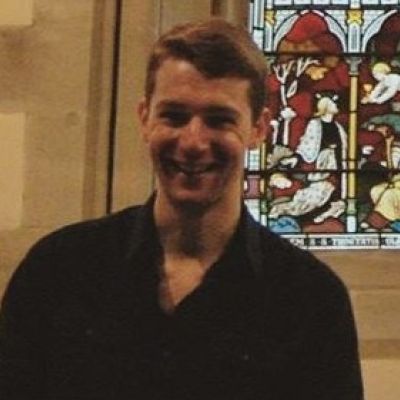 A head and shoulders shot of a smiling Peter Waterman stood in front of a stained glass window wearing a dark coloured shirt