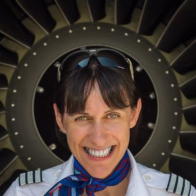 Head and shoulders shot of a smiling Sarah Barry stood in front of an airline jet engine in a pilots uniform