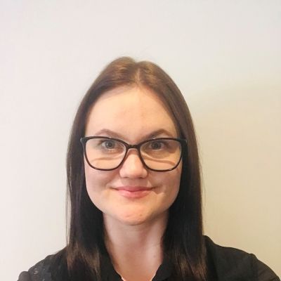 Head and shoulders shot of a smiling Taryn Tavener Smith wearing glasses stood facing into the camera in front of a white wall
