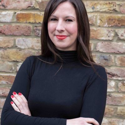 A head and shoulders shot of Lara Baker stood with her arms crossed wearing a black jumper and looking directly into the camera