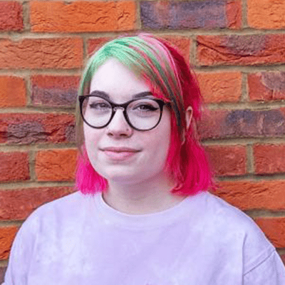 A headshot of a smiling Maddy Kendal-Murray wearing glasses and stood in front of a brick wall