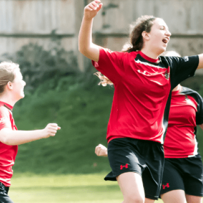 Three women from the BNU football team celebrating on the football pitch, wearing their red and blue kit