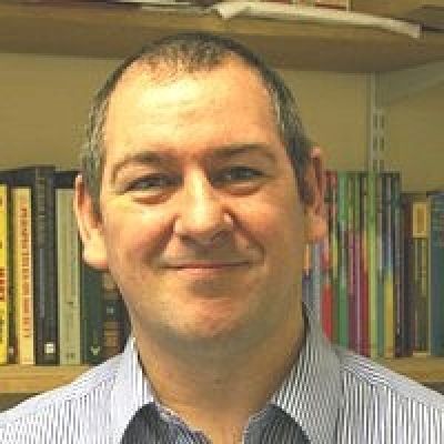 Headshot of a smiling Des McDermott stood in front of a book case wearing a shirt, smiling directly into the camera