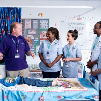 Four student nurses in uniform stood around a simulation hospital bed with a child laying down on it