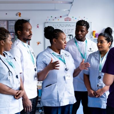 Five student nurses in uniform stood in discussion whilst facing a lecturer in a simulation ward