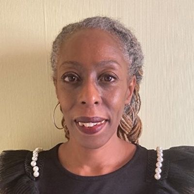 A head and shoulders photo of a smiling Brenda Brown wearing a black dress stood against a grey wall looking directly into the camera