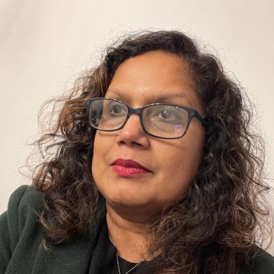 Photo of Sharmin, who has light brown skin, long curly brown hair, glasses, and is wearing a black jacket over a dark shirt.