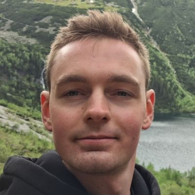A headshot of Kamil Bilski wearing a black hoodie standing in front of a mountain and lake.