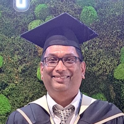 Headshot of Sunny Daby, wearing a graduation gown and hat against a green wall.