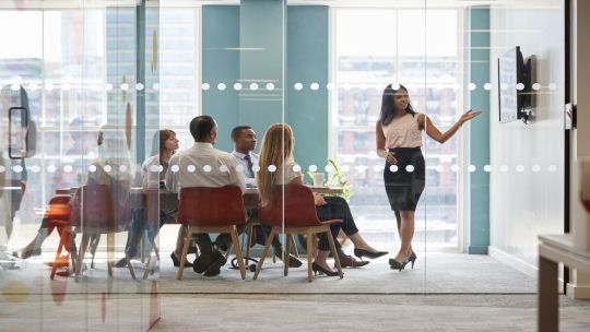 A lady stood up showing a presentation with 6 others sat at a desk watching