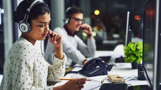 Female on headset phone writing down notes. Man in background on phone